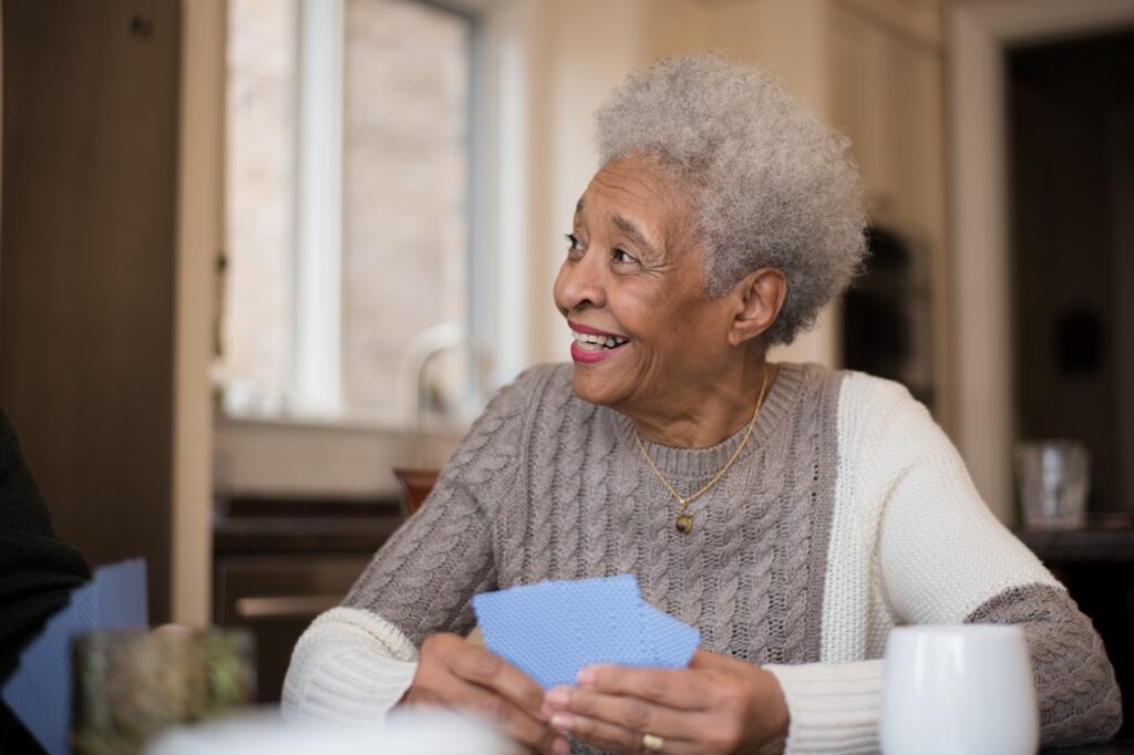 A senior woman of African descent smiles as she plays cards with friends off camera. She is also sitting at a kitchen table drinking coffee.